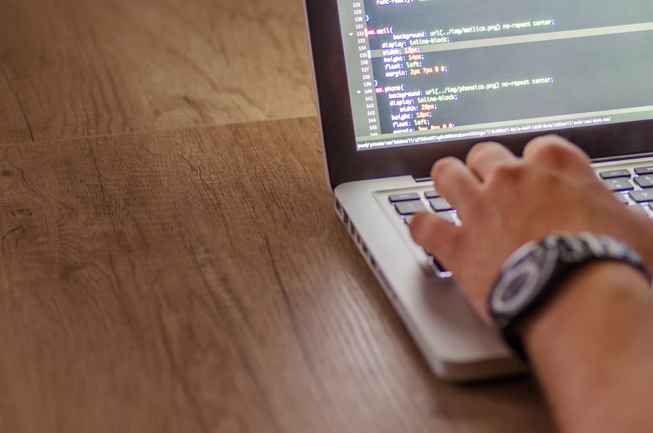 A person working on a laptop placed on a wooden desk, with the screen displaying lines of programming code in a text editor. The individual's right hand is resting on the keyboard, wearing a wristwatc
