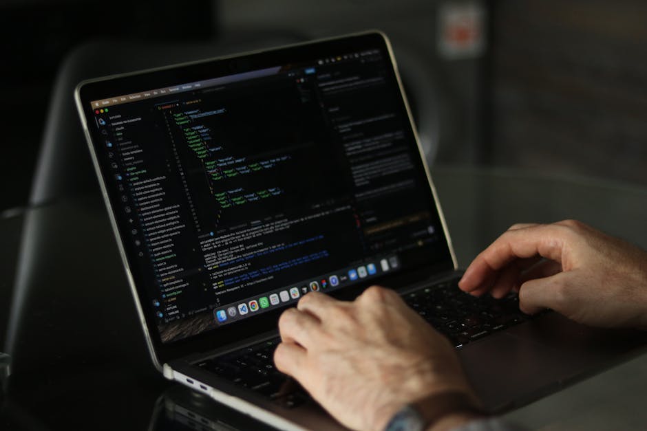 A person working on a laptop placed on a wooden desk, with the screen displaying lines of programming code in a text editor. The individual's right hand is resting on the keyboard, wearing a wristwatc